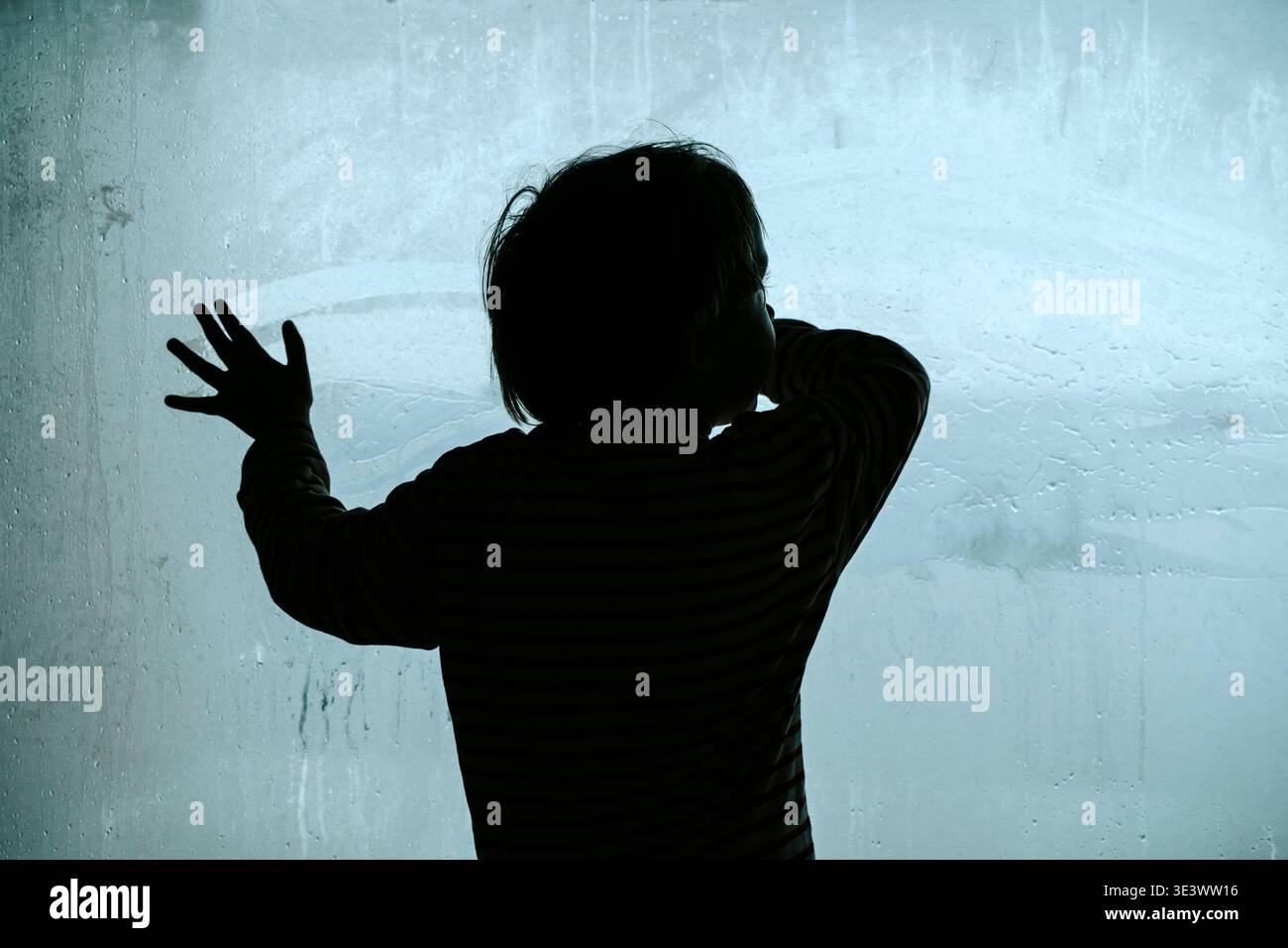 Silhouette d'un enfant solitaire regardant à travers une grande fenêtre embuée froide. Un travail de photographie conceptuelle explorant les thèmes de la dépression infantile, de la crise psychologique et du profond sentiment d’isolement et de tristesse. Photo de haute qualité Banque D'Images