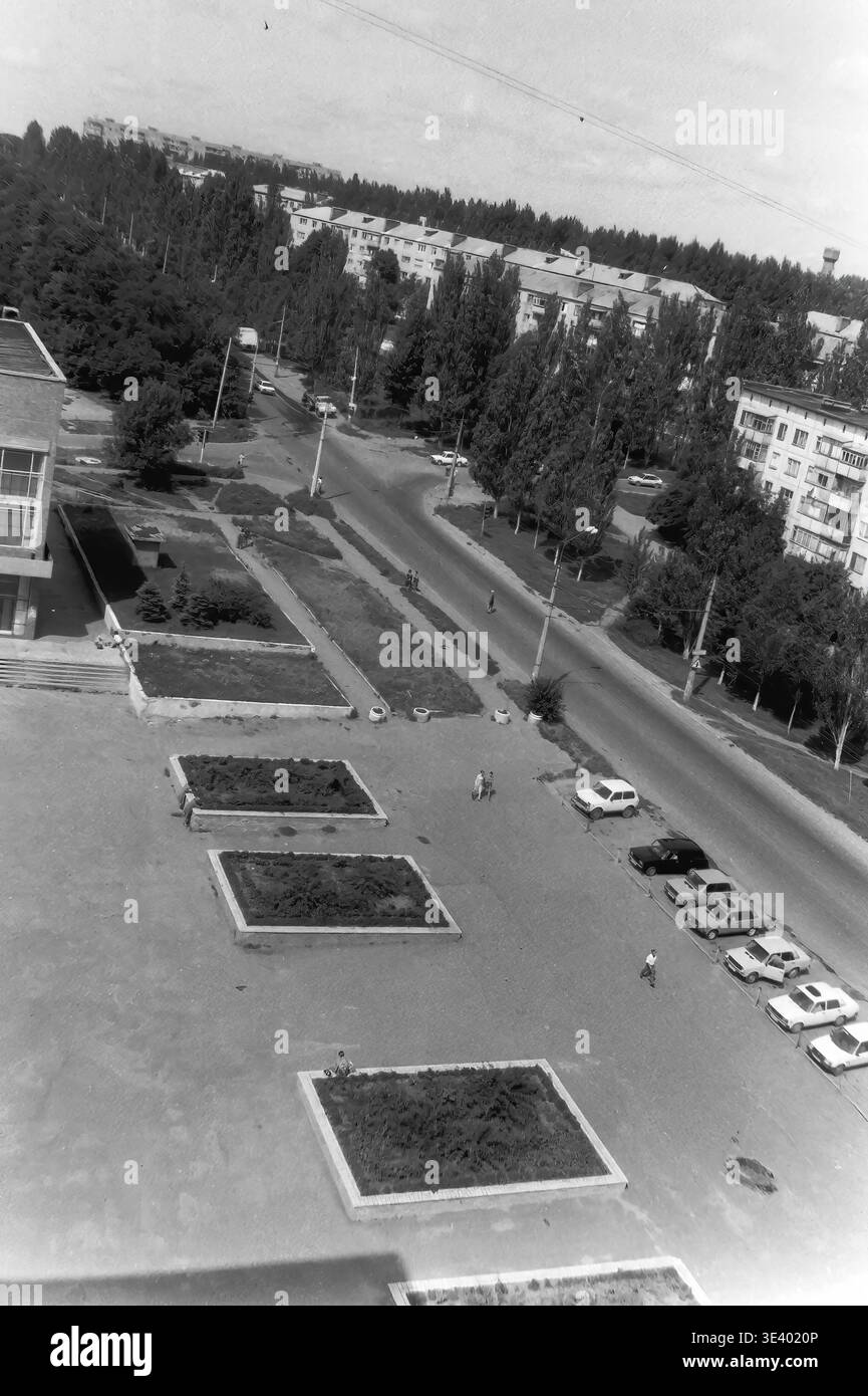 Sloviansk, milieu des années 1990, Artem Microdistrict (zone de l'Institut pédagogique). Une photographie historique en noir et blanc montrant une vue impressionnante sur une place municipale spacieuse. La zone pavée présente plusieurs parterres de fleurs rectangulaires, avec une rangée de diverses voitures domestiques garées soigneusement sur le côté droit. Les citoyens locaux sont vus marchant tranquillement à travers la place ouverte vers la large avenue bordée d'arbres de la ville. Cet instantané d'archives exceptionnellement détaillé capture magnifiquement l'infrastructure post-soviétique et la vie dans le Donbass paisible. Banque D'Images