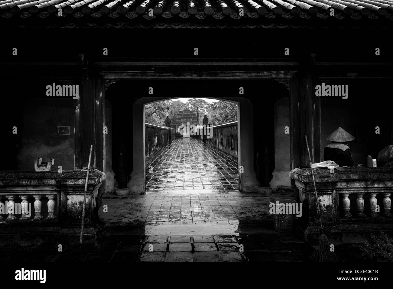 Passerelle imbibée de pluie à travers une porte sombre encadrant une arche lointaine avec un ouvrier dans un chapeau conique, ville impériale de Hué, Vietnam. Banque D'Images