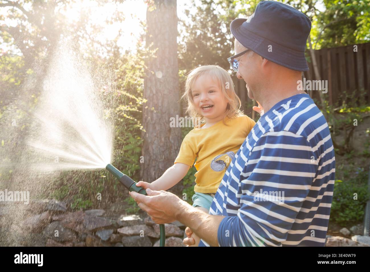 Père tenant son rire heureux fils de 2 ans tout en arrosant les plantes dans le jardin par une journée d'été ensoleillée. Bonheur familial et joie extérieure. Banque D'Images