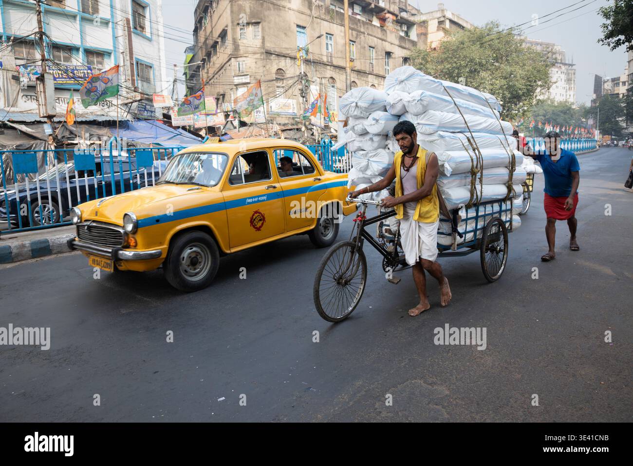 Un taxi jaune emblématique ambassadeur de l'Hindustan passant devant un homme poussant un rickshaw van, Kolkata, Bengale occidental, Inde Banque D'Images