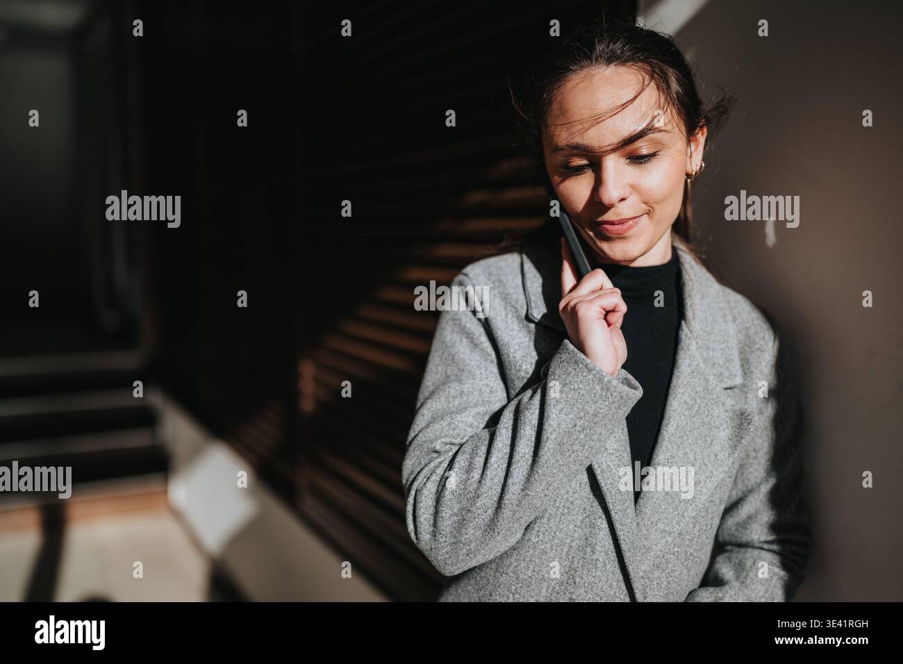 Femme d'affaires au téléphone parlant avec une équipe tout en s'appuyant contre un mur ensoleillé dans un manteau Banque D'Images
