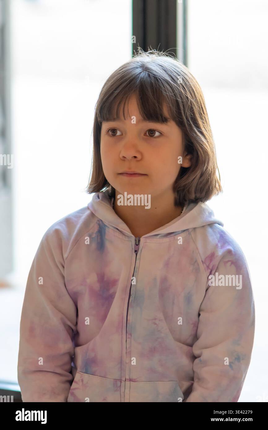 Fille d'environ 8 ans est assis près de la fenêtre et regarde de côté. Banque D'Images