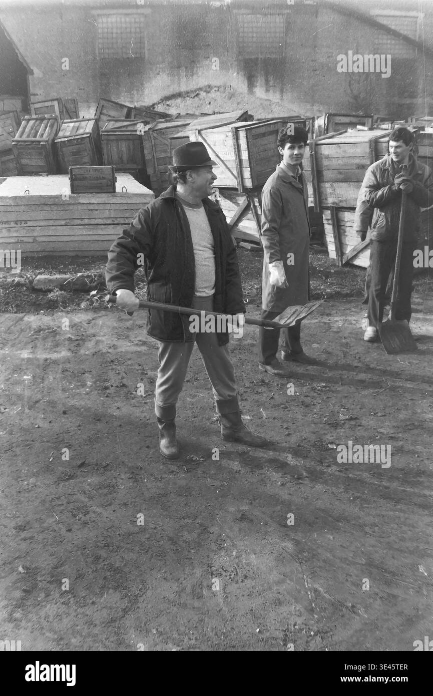 Sloviansk, avril 1989, cour d'usine AIZ. Un directeur d'usine joyeux portant un chapeau classique tient fièrement une lourde pelle horizontalement comme une arme pendant un bref moment de repos lors d'un événement traditionnel de nettoyage de volontaires subbotnik de printemps. Il sourit chaleureusement tout en étant entouré de ses collègues observateurs dans la cour industrielle remplie de caisses en bois. Ce portrait franc reflète merveilleusement l’humeur positive, l’humour et la forte camaraderie du collectif de travail dans le Donbass paisible. Banque D'Images