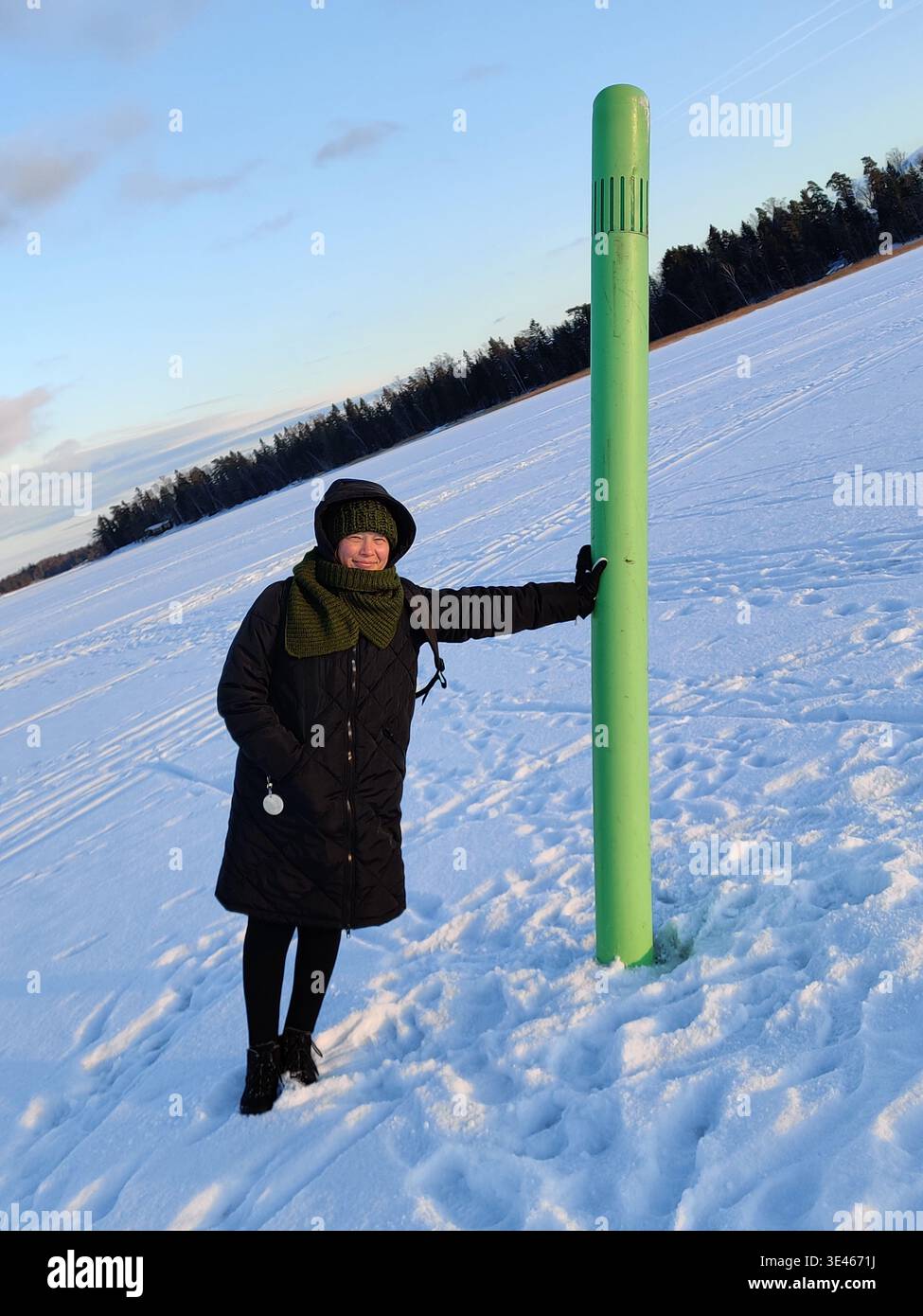 Une femme contente, vêtue d'un manteau d'hiver noir, d'un bonnet tricoté vert et d'une écharpe, pose avec un sourire chaleureux tandis qu'elle s'appuie contre une grande marque vert vif Banque D'Images