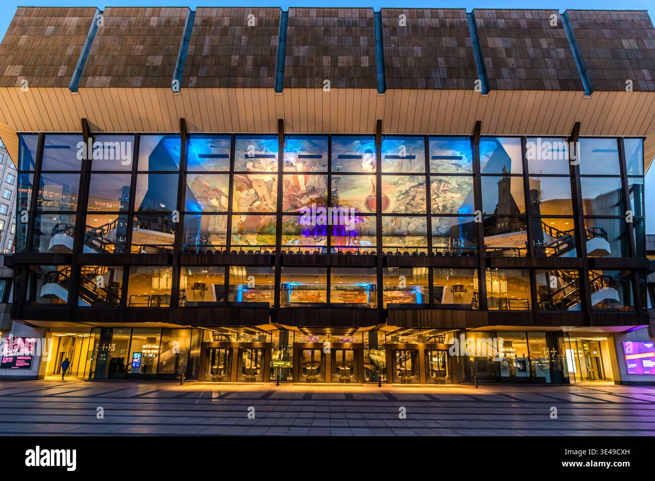 Gewandhaus de Leipzig au crépuscule : la façade vitrée illuminée de la salle de concert révèle la peinture à grande échelle du plafond dans le hall ; la zone d’entrée est parsemée de quelques visiteurs. Leipzig, Saxe, Allemagne Banque D'Images