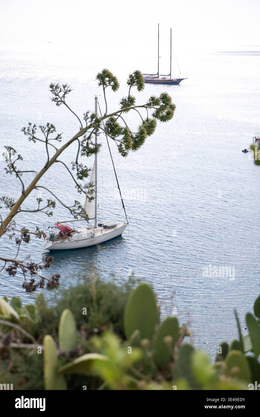 Vue d'un voilier blanc ancré dans les eaux bleues calmes de la mer Méditerranée encadrée par la végétation côtière à Lipari, Sicile, Italie. Banque D'Images