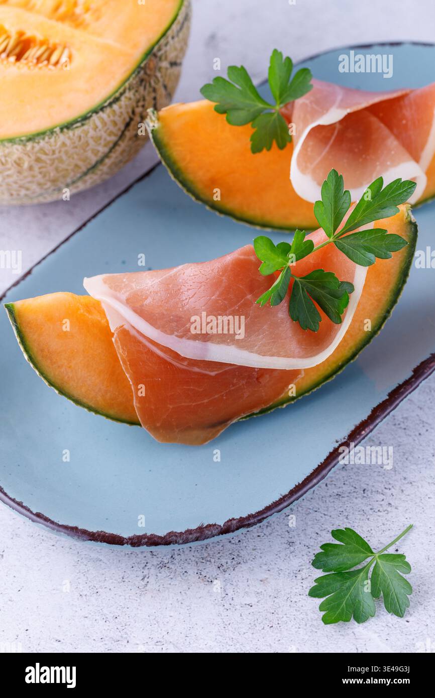 Tranches de jambon espagnol traditionnel (viande de porc séchée) avec melon (cucumis melo) et feuilles de persil frais herbes sur assiette vintage. Plat sur table de pierre légère Banque D'Images