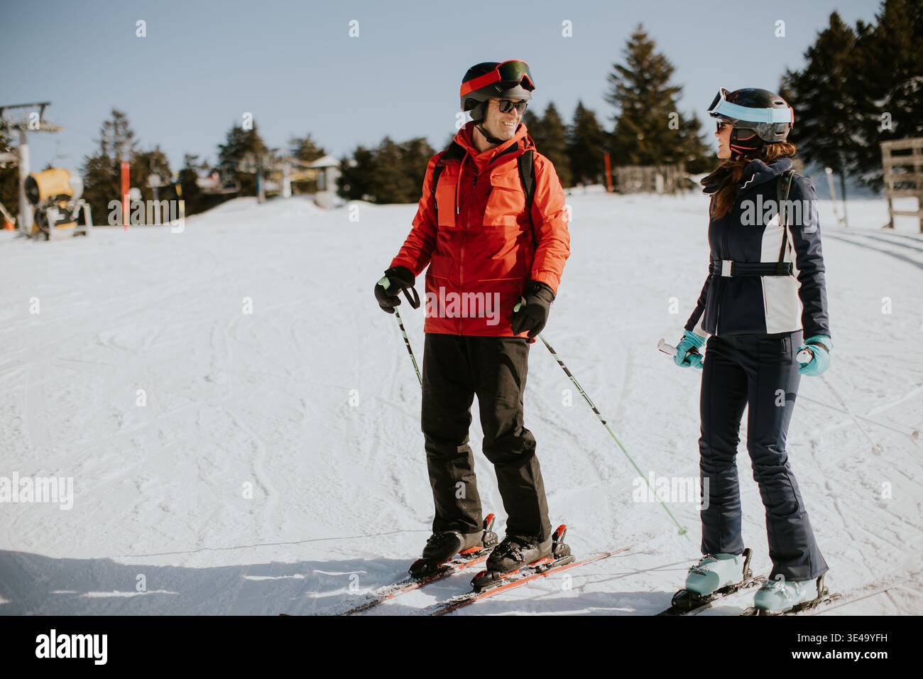 Deux amis se tiennent debout sur des skis dans une station de ski enneigée. Ils discutent et profitent du ciel bleu et du soleil pendant leur sortie hivernale. A proximité, remontées mécaniques et arbres Banque D'Images