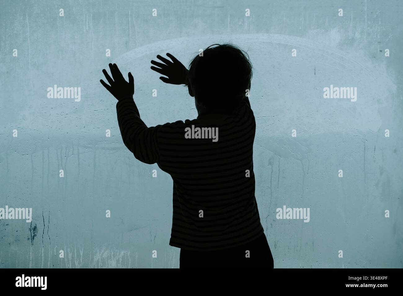 Silhouette d'un enfant solitaire regardant à travers une grande fenêtre embuée froide. Un travail de photographie conceptuelle explorant les thèmes de la dépression infantile, de la crise psychologique et du profond sentiment d’isolement et de tristesse. Photo de haute qualité Banque D'Images