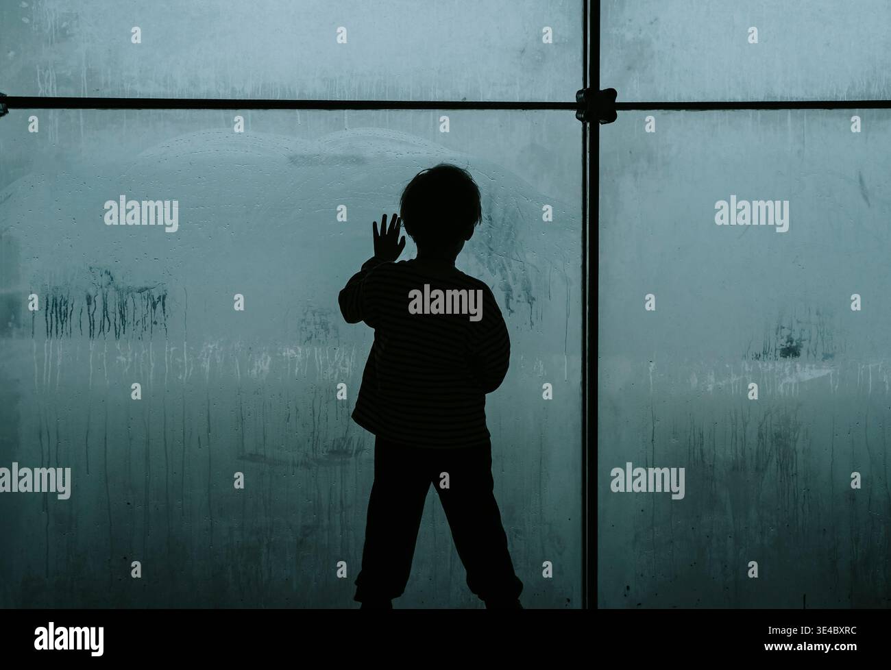 Silhouette d'un enfant solitaire regardant à travers une grande fenêtre embuée froide. Un travail de photographie conceptuelle explorant les thèmes de la dépression infantile, de la crise psychologique et du profond sentiment d’isolement et de tristesse. Photo de haute qualité Banque D'Images