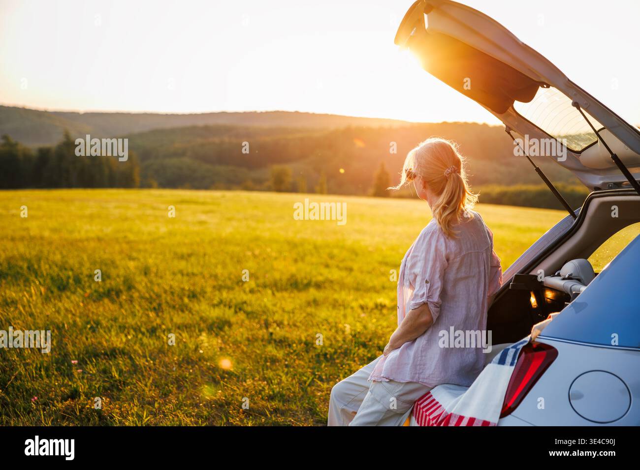 Femme assise sur le coffre de voiture appréciant le coucher du soleil pendant le voyage en solo. Touriste féminine se relaxant pendant le wanderlust sur le voyage de vacances d'été Banque D'Images