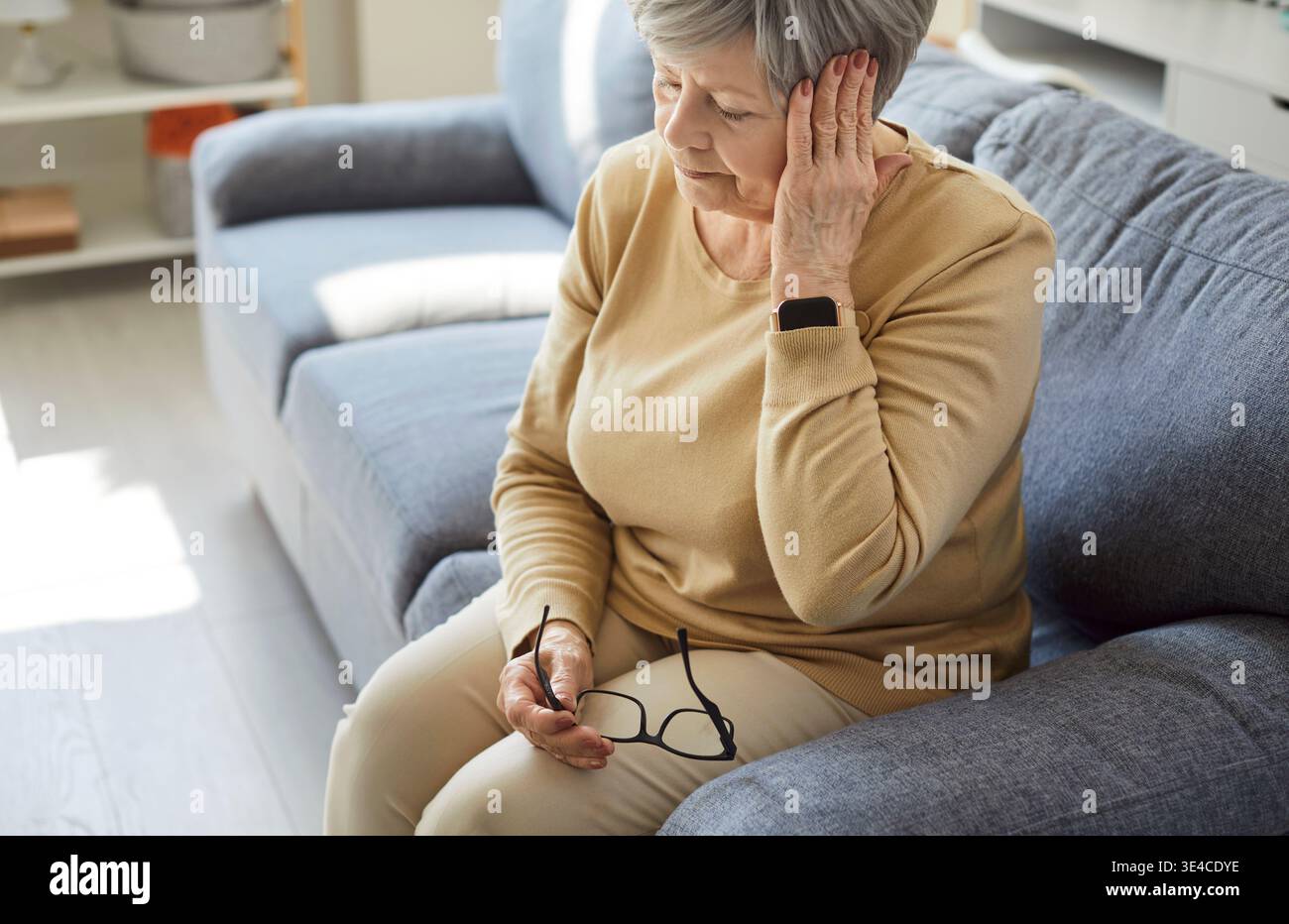 Femme âgée triste fatiguée avec mal de tête assise sur le canapé à la maison, souffrant de douleur Banque D'Images