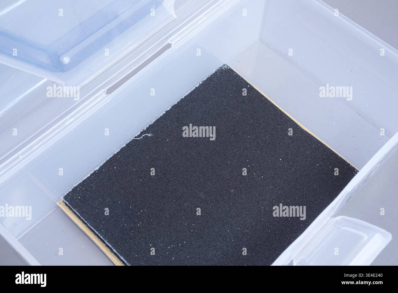 Le papier abrasif noir repose dans un récipient en plastique transparent. La surface lisse montre une texture fine et abrasive. Idéal pour le lissage du bois, du métal ou du plastique. Propre, m Banque D'Images