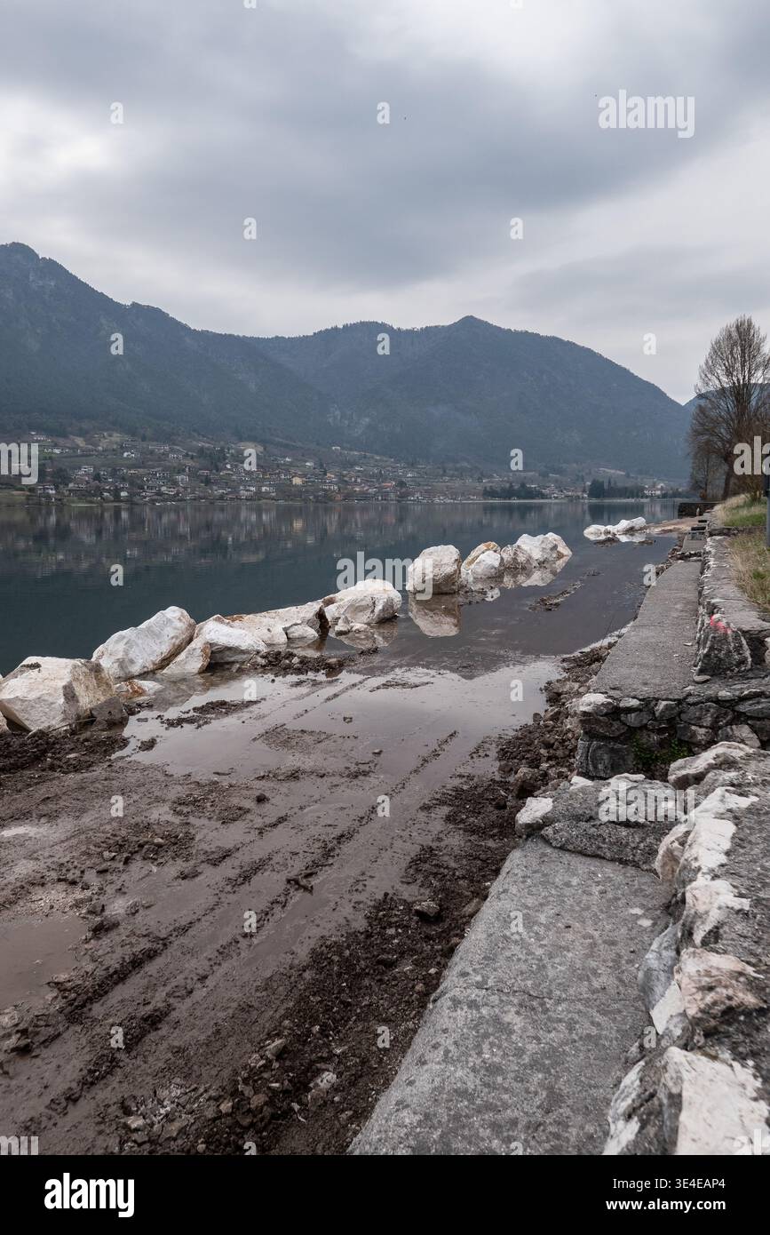 Travaux de construction de pistes cyclables le long de la rive du lac Idro avec de grandes pierres protectrices terrain boueux et reflets de montagne dans le paysage d'eau calme SC Banque D'Images