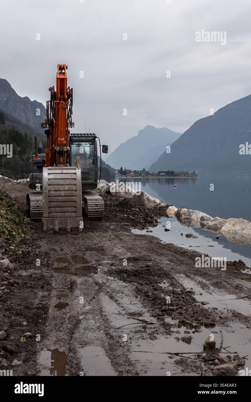 Excavatrice opérant sur la construction de pistes cyclables le long de la rive du lac Idro avec des pierres de protection de sol boueux et une infrastructure de paysage de montagne pro Banque D'Images