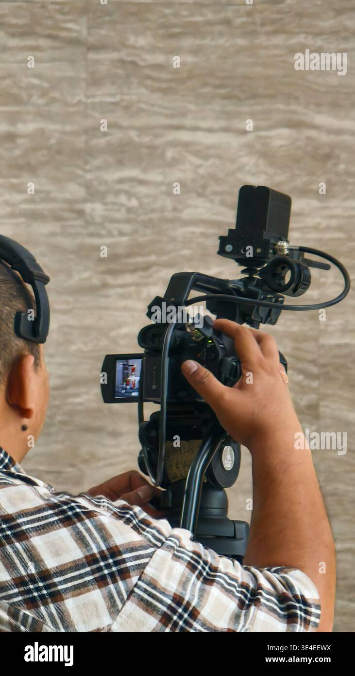 Personne utilisant une caméra vidéo professionnelle sur trépied, portant une chemise à carreaux et un casque, ajustant les paramètres de la caméra à l'intérieur. Banque D'Images