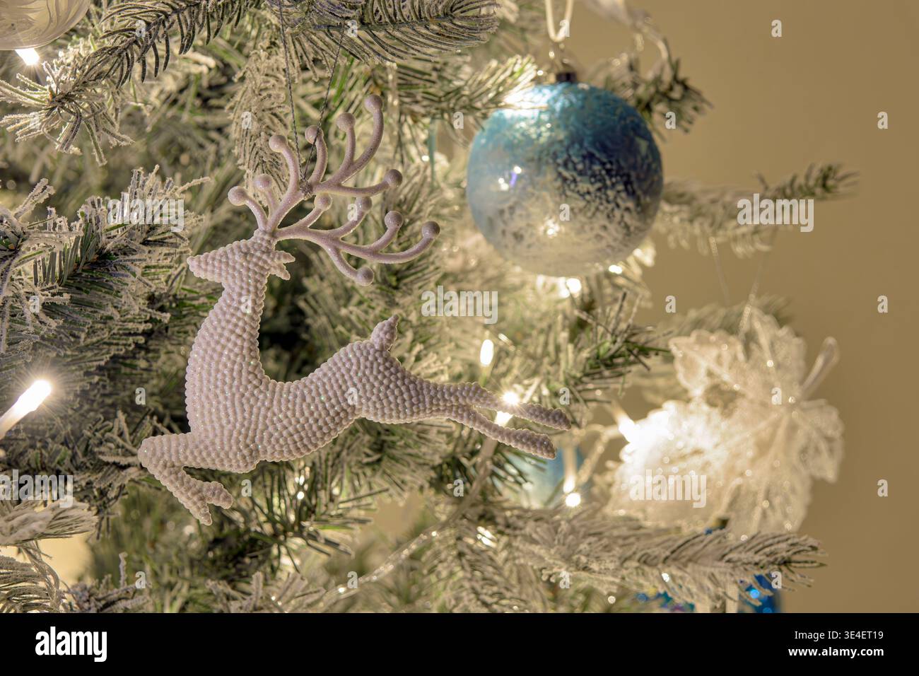 Gros plan d'un renne scintillant suspendu à un sapin blanc floqué avec boule et lumières bleues. Banque D'Images