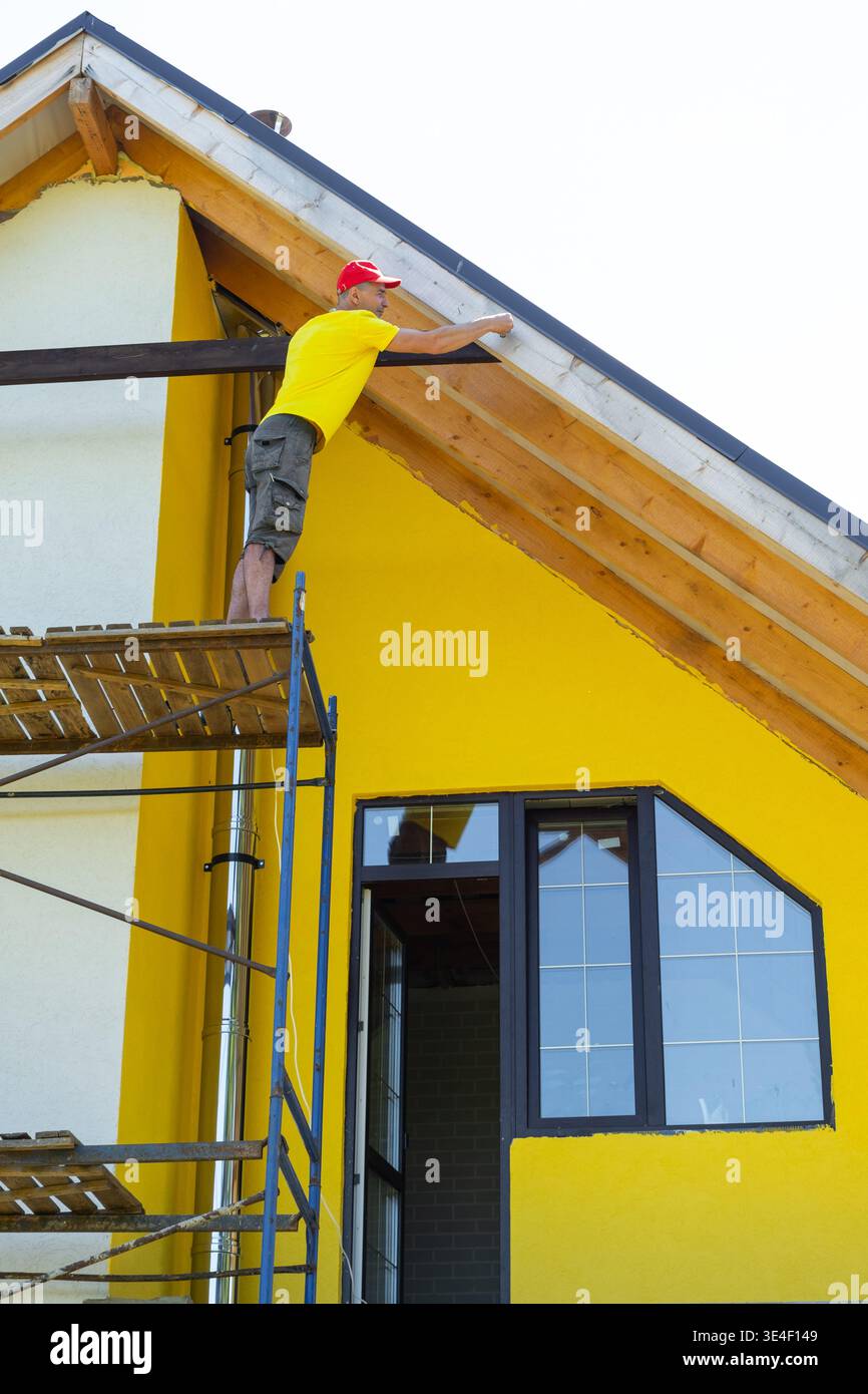 Équilibre ouvrier sur l'échafaudage supérieur fixant la planche horizontale sous le pignon, façade contemporaine jaune, moment critique de construction, sécurité personnelle GE Banque D'Images