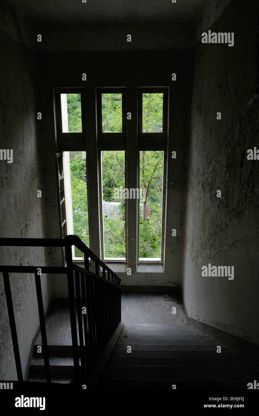 Grandes fenêtres éclairant des escaliers sombres et poussiéreux, jeux d'ombre et de lumière dans une maison abandonnée, décor d'urbex inquiétant Banque D'Images