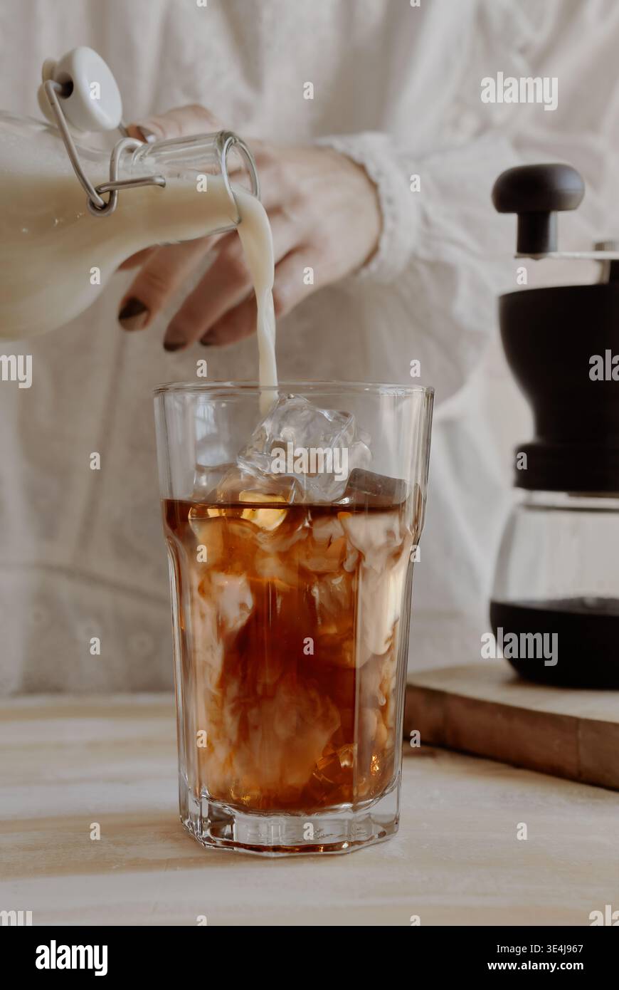 Gros plan des mains versant du lait dans un verre de café glacé avec des glaçons, créant un tourbillon crémeux sur un comptoir léger à côté d'une carafe à infusion froide dans un c. Banque D'Images