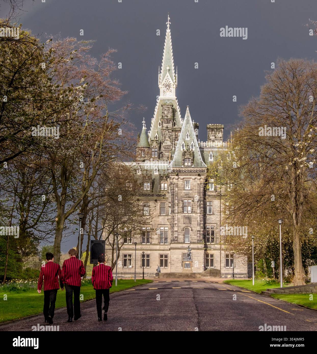 Trois étudiants en blazers rouges marchent le long d'une route bordée d'arbres vers le bâtiment principal orné de style néo-gothique du Fettes College à Édimbourg, évoquant Trad Banque D'Images