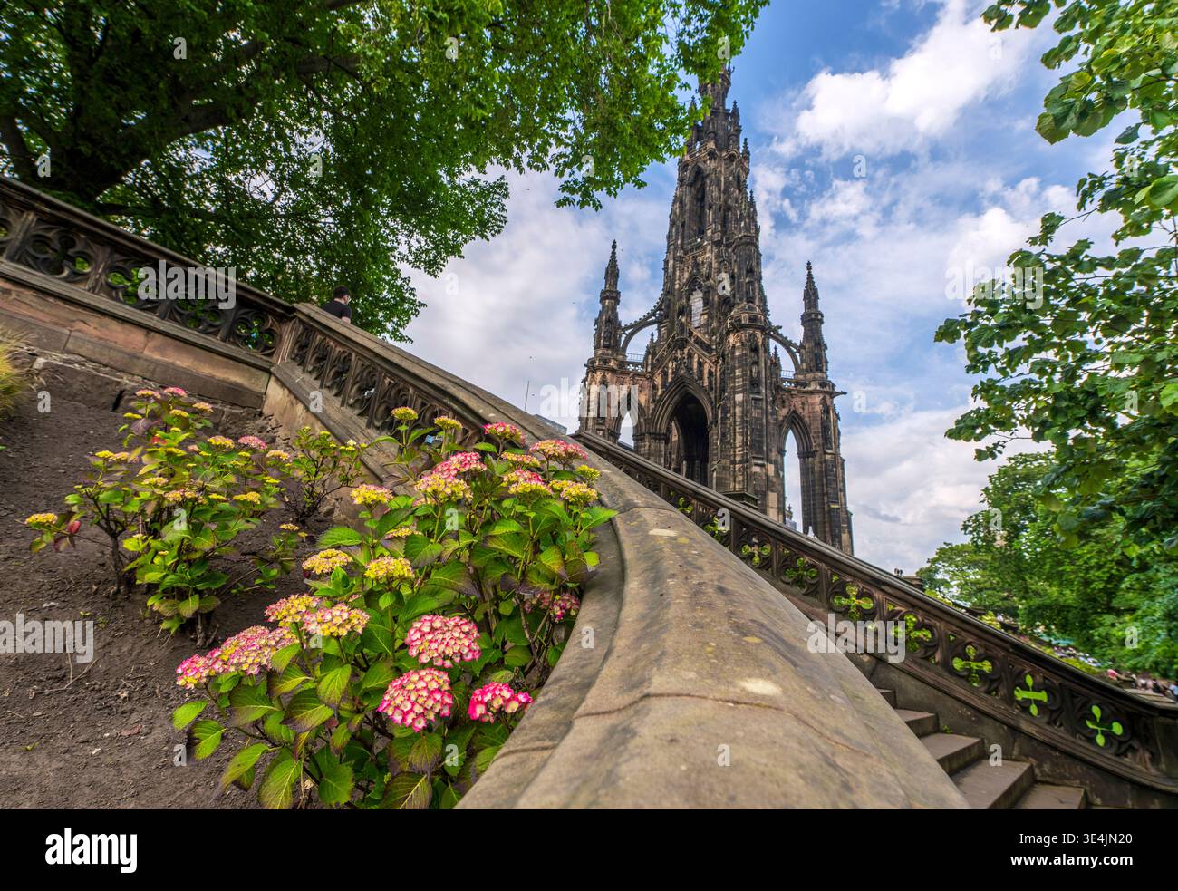 Vue spectaculaire en bas angle du Scott Monument encadré par des arbres feuillus, des escaliers en pierre et des hortensias en fleurs. L'architecture gothique historique contraste avec Banque D'Images