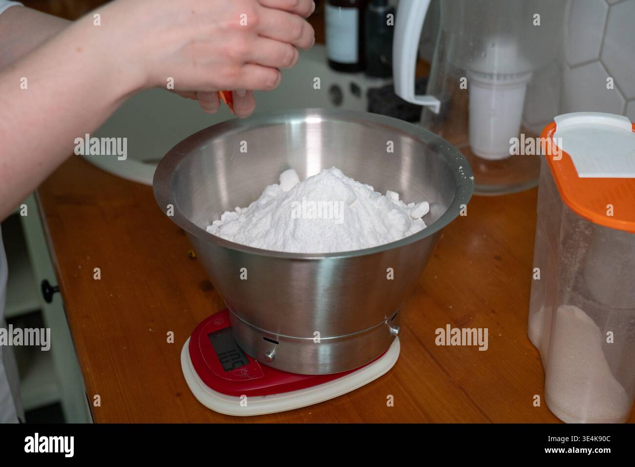 Main ajoutant l'ingrédient dans la farine dans un bol en métal sur l'échelle de cuisine, préparation de cuisson sur le comptoir en bois dans la cuisine à la maison. Contenu de cuisson, recette Banque D'Images