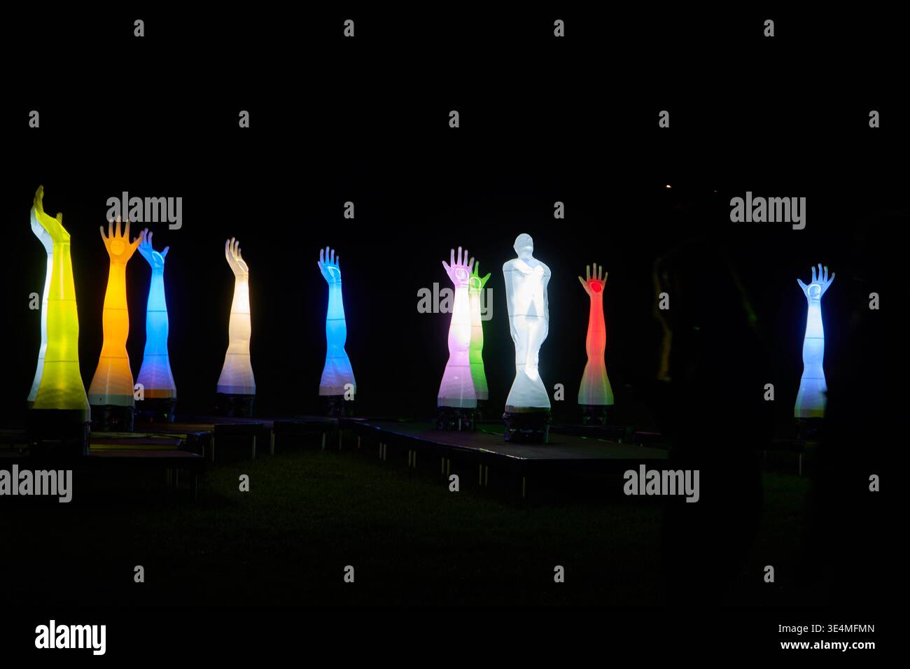 Une installation d'art nocturne en plein air avec de multiples sculptures illuminées de bras et de mains humains s'étendant vers le haut, affichées dans des couleurs vives Banque D'Images
