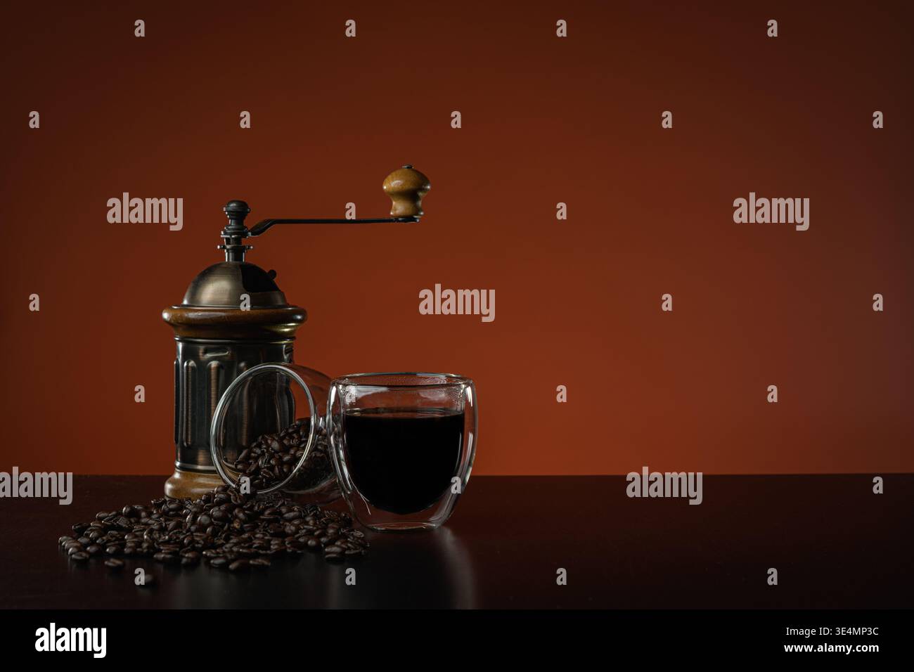Une boisson chaude au café dans une tasse en verre à double paroi sur une table en bois brun foncé, à côté d'un moulin manuel et d'une autre tasse avec des grains de café débordant. Banque D'Images