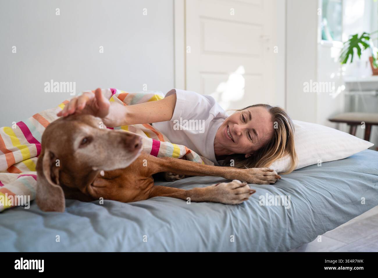 Souriante femme détendue câlin avec chien Vizsla au lit le matin, partageant des moments joyeux avec animal de compagnie. Banque D'Images