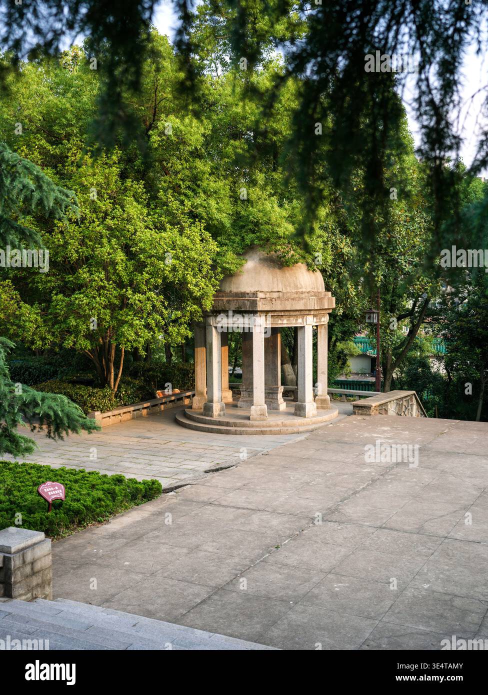 Petit pavillon circulaire blanc en pierre avec colonnes classiques, situé sur une plate-forme pavée entourée d'arbres verdoyants dans un parc public. La scène Banque D'Images
