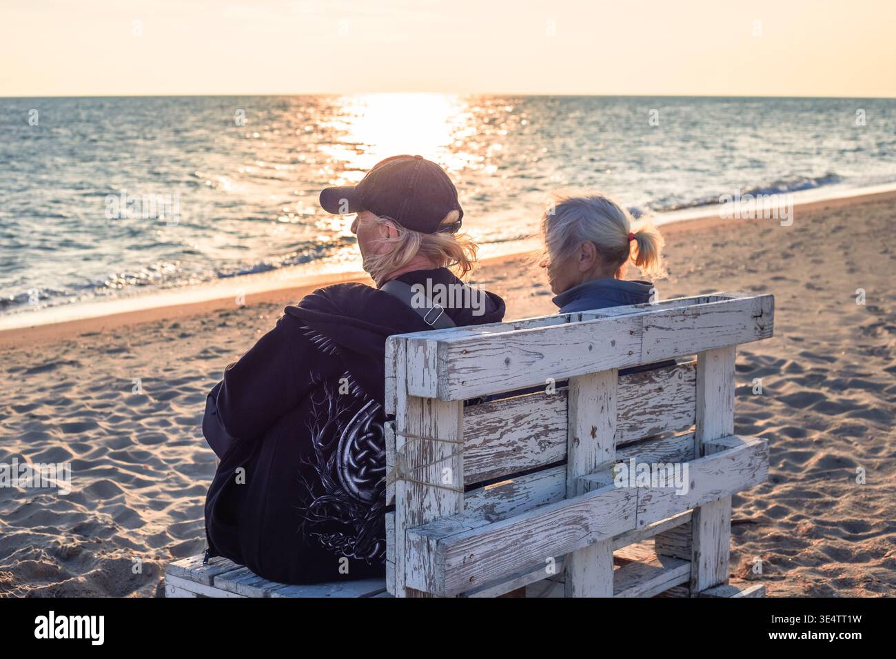 Un homme et une vieille femme sont assis sur un banc en bois sur une plage de sable fin regardant le coucher de soleil sur l'eau. père et fils, vacances à la plage, vue sur le coucher du soleil. Banque D'Images