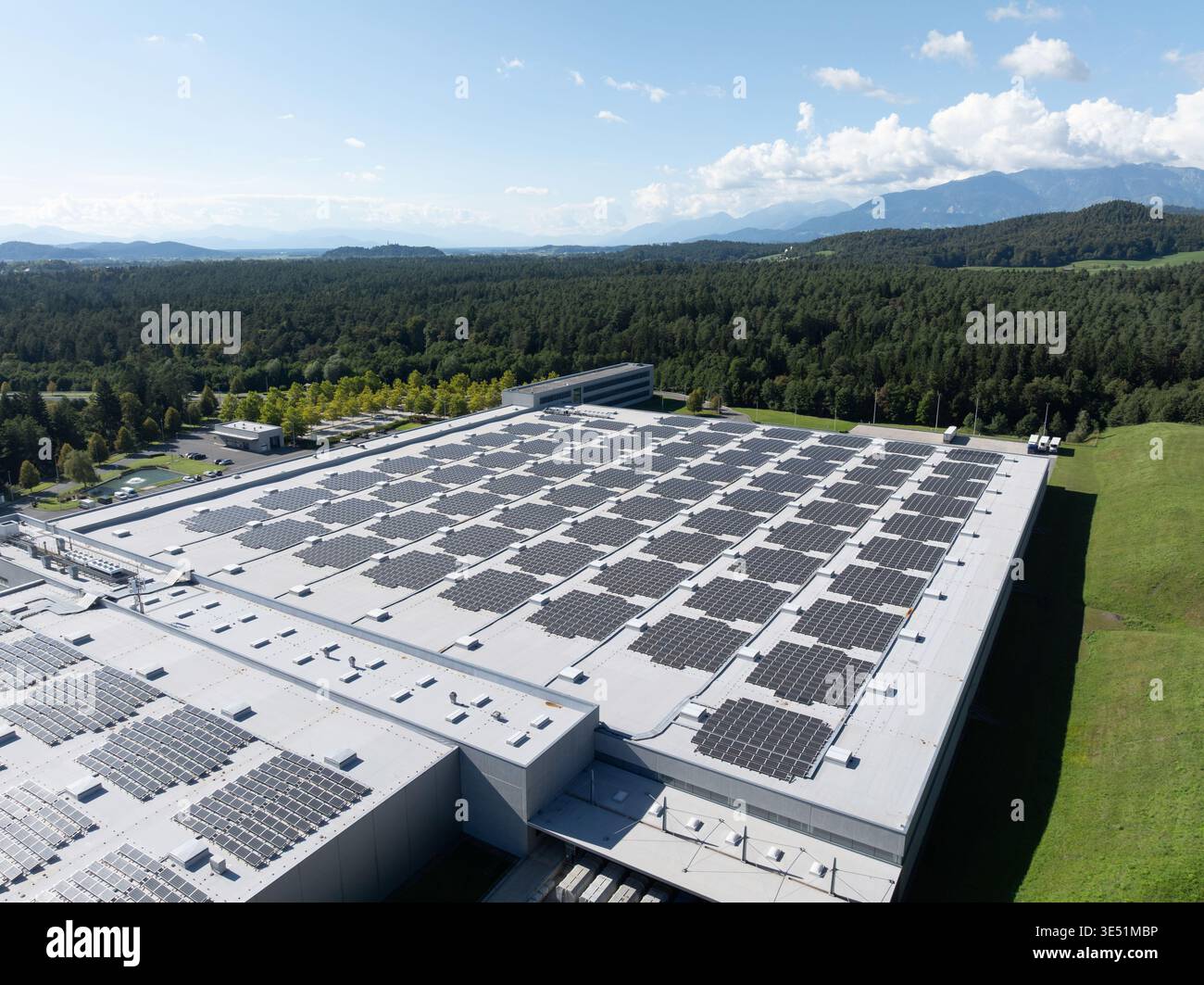Vue aérienne d'un centre logistique montrant des entrepôts, des quais de chargement et des camions entourés de champs verdoyants et de montagnes. Banque D'Images