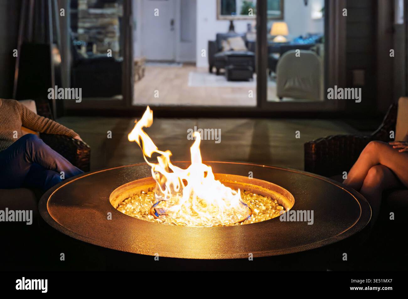 Un jeune couple s'assoit près d'un foyer flamboyant sur une terrasse de jardin extérieure avec une pièce éclairée derrière eux la nuit. Banque D'Images