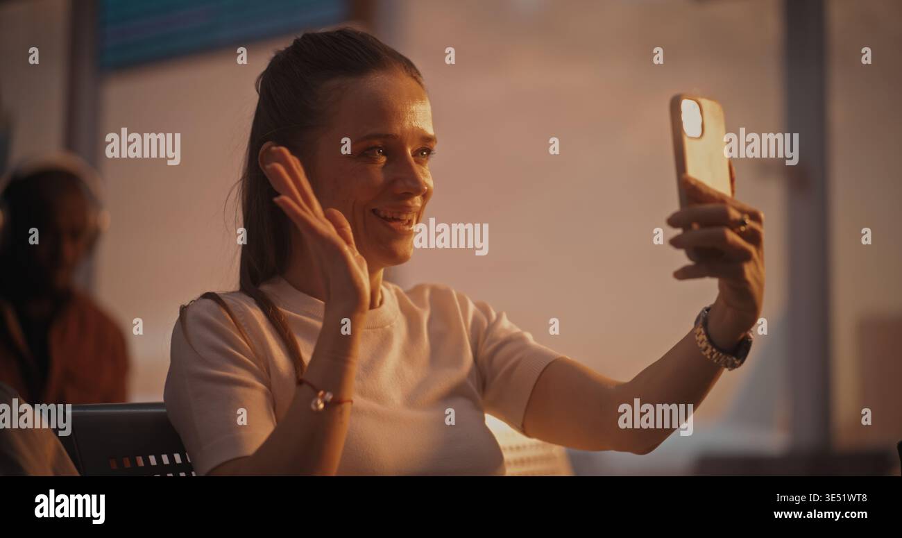 Assise avec la valise près, la femme s'engage dans un appel vidéo pendant l'escale. Femme regarde dans la caméra du téléphone, riant et discutant. Mode de vie des voyageurs connectés. Communications mobiles, connectivité 5G. Banque D'Images