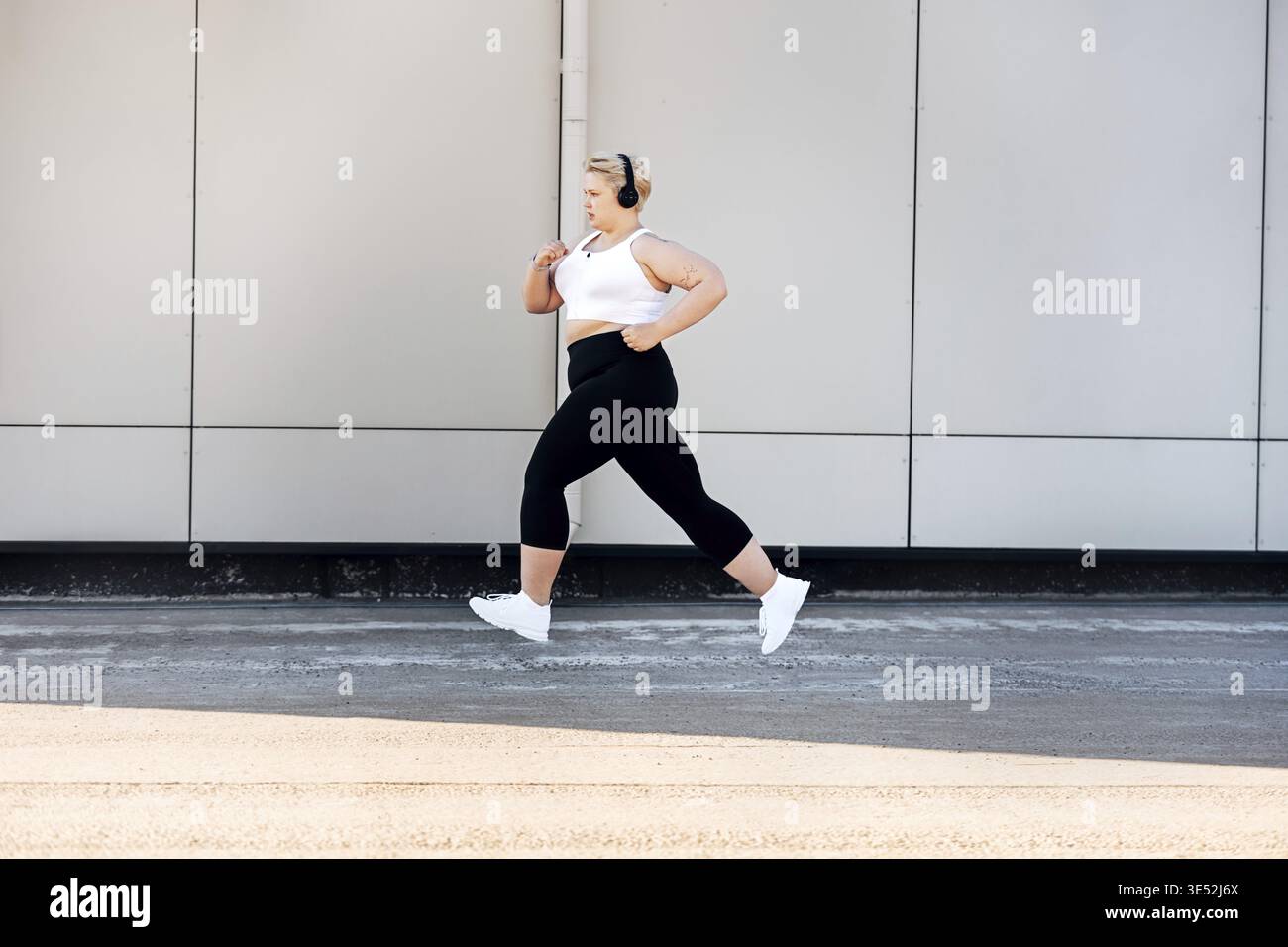 Vue latérale d'une femme grande taille portant des écouteurs près d'un mur. Femme courbée dans des vêtements de sport jogging à l'extérieur Banque D'Images