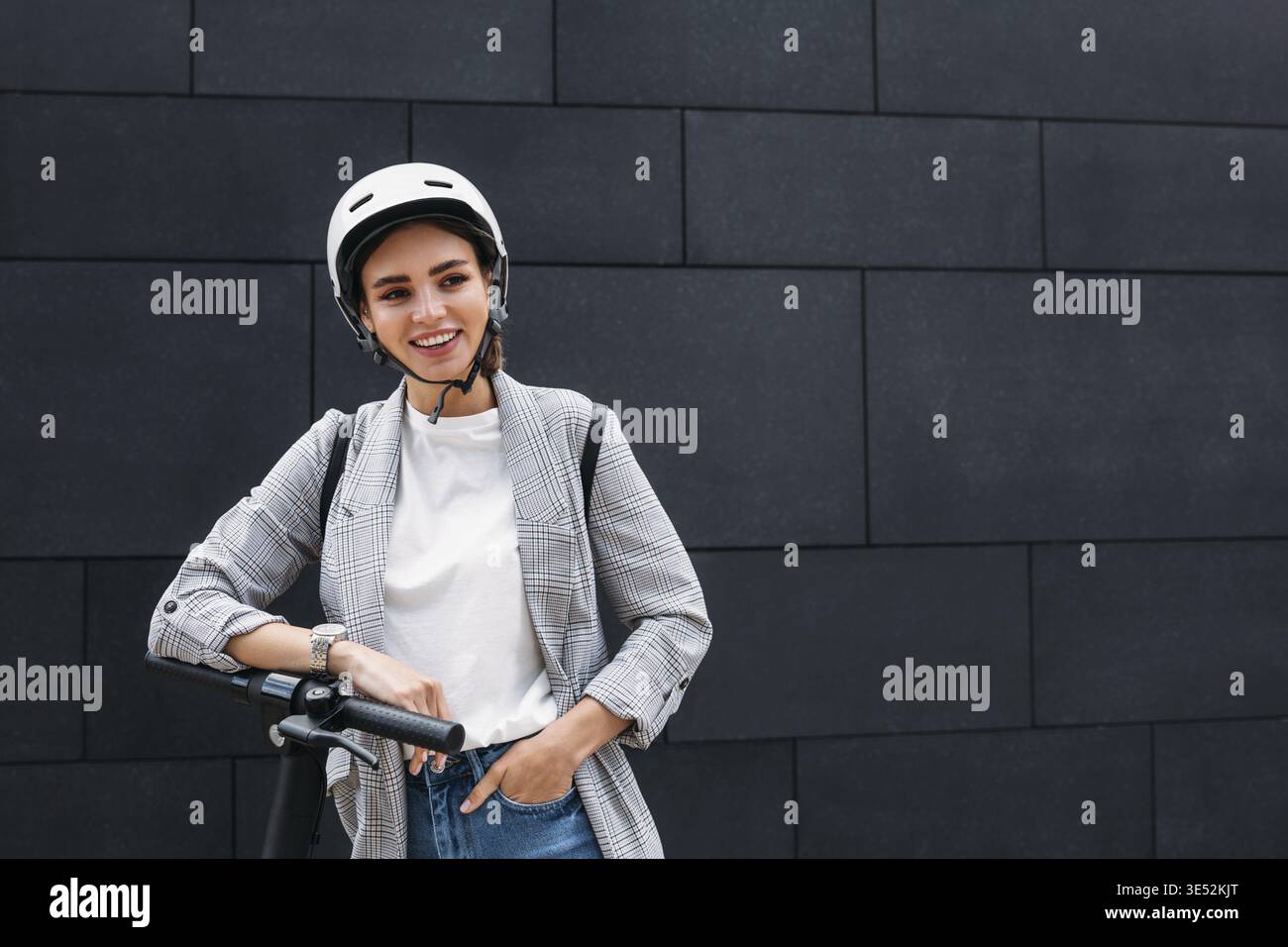 Portrait d'une femme d'affaires souriante penchée sur un guidon de scooter électrique. Jeune femme gaie dans un casque de vélo au mur noir qui regarde loin Banque D'Images