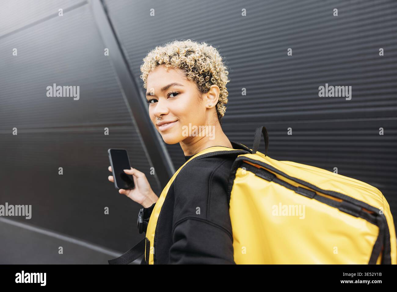 Portrait d'une femme de livraison souriante avec un sac debout sur un trottoir et tenant un smartphone Banque D'Images