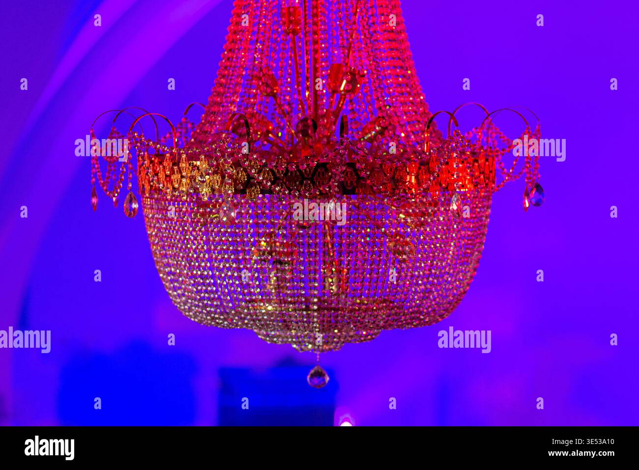 Vue rapprochée d'un lustre suspendu avec des brins de cristal et des prismes, brillant dans des tons rouges sur un fond violet éclairé pour une installation de salle ou de scène. Banque D'Images