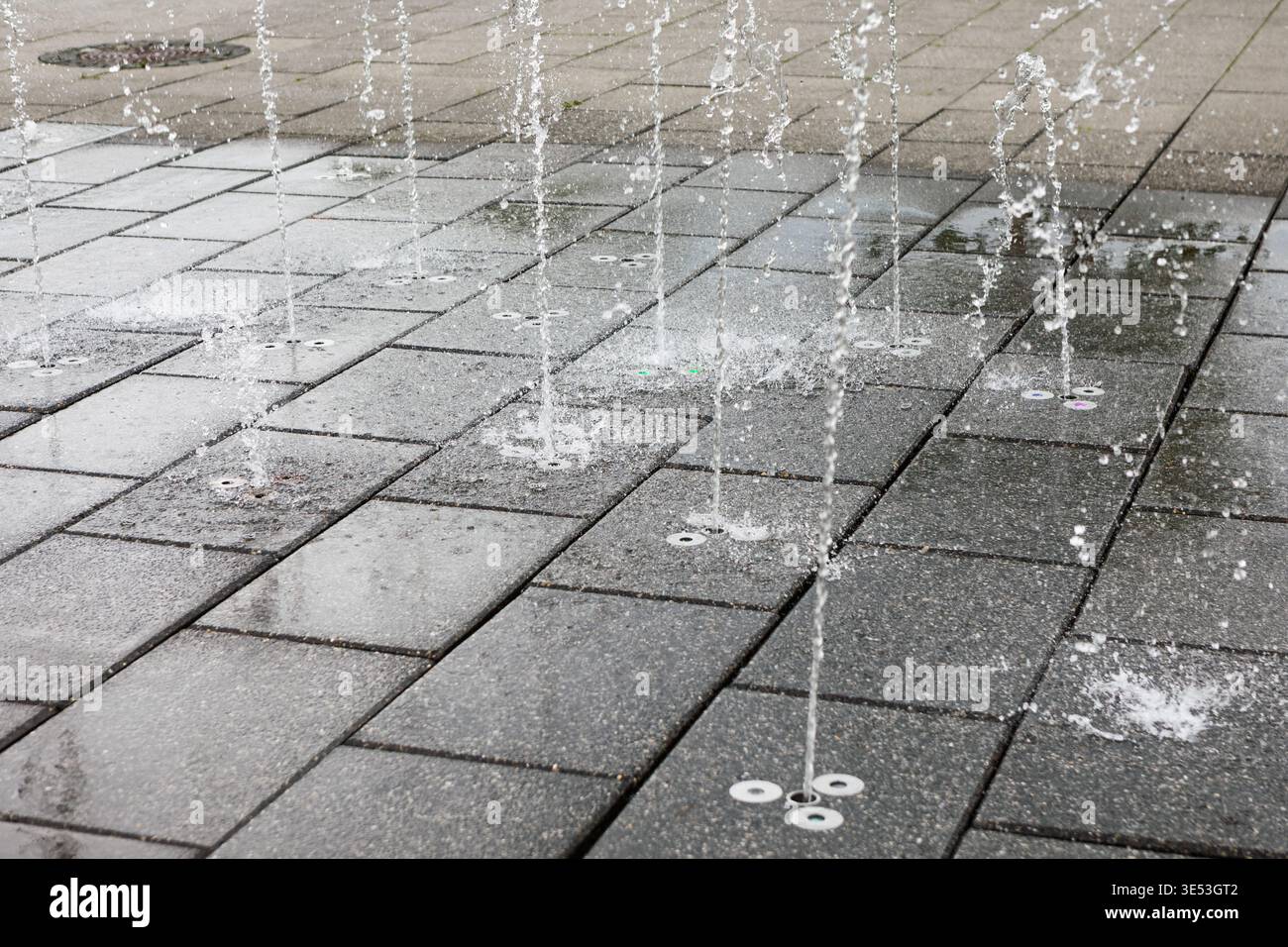 Des jets d'eau projettent vers le haut depuis le sol dans une fontaine moderne, créant des ruisseaux ludiques sur des carreaux de pierre gris. La conception géométrique propre ajoute une réf Banque D'Images