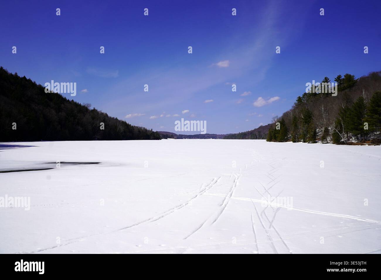 Une vaste étendue enneigée de lac Philippe gelé est encadrée par des collines densément boisées sous un ciel bleu vif dans le parc de la Gatineau, au Québec Banque D'Images