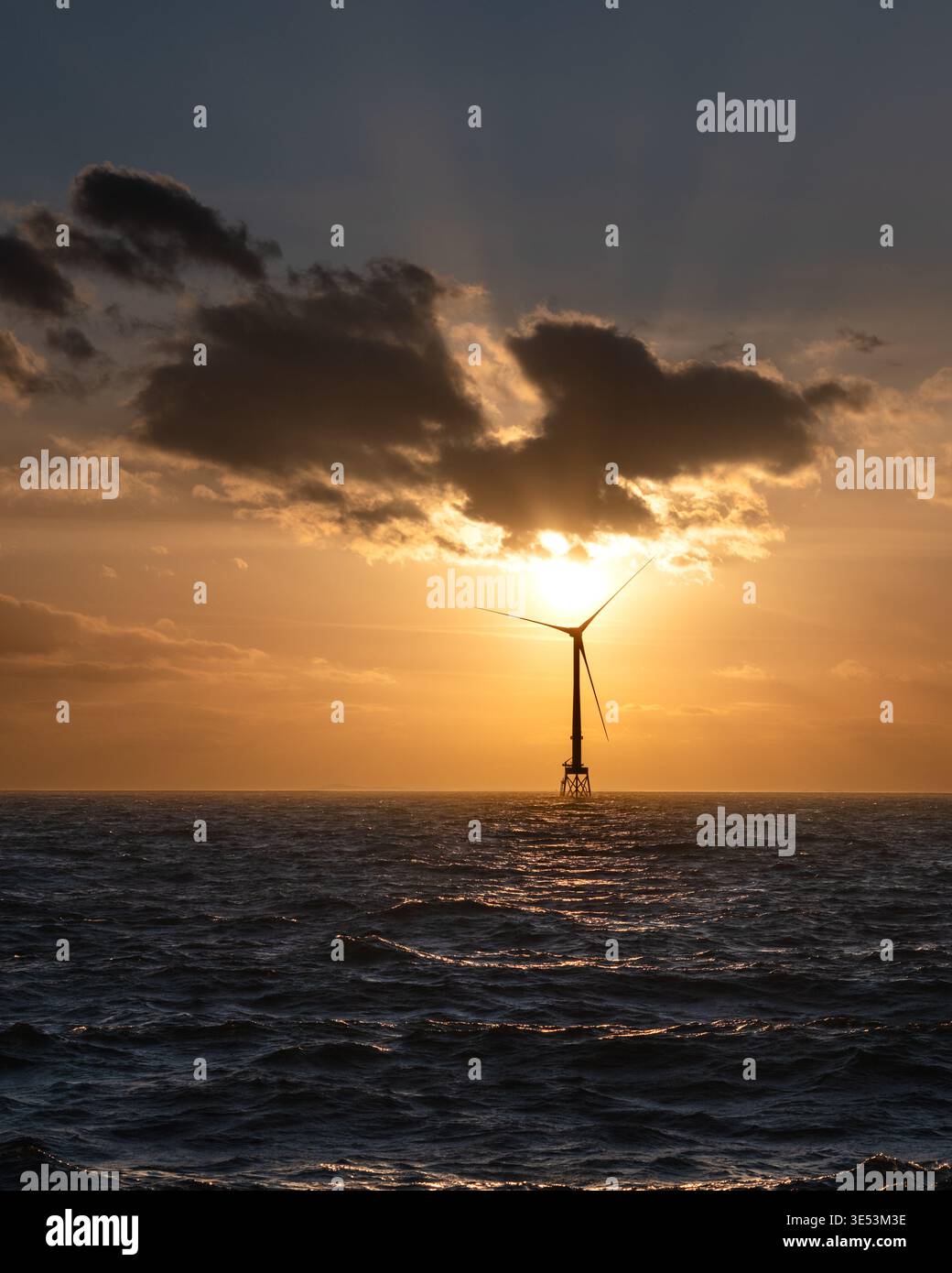 Des éoliennes offshore se sont dessinées contre un coucher de soleil ambré flamboyant sur la mer du Nord, des rayons crépusculaires traversant les nuages derrière l'éolienne. Banque D'Images