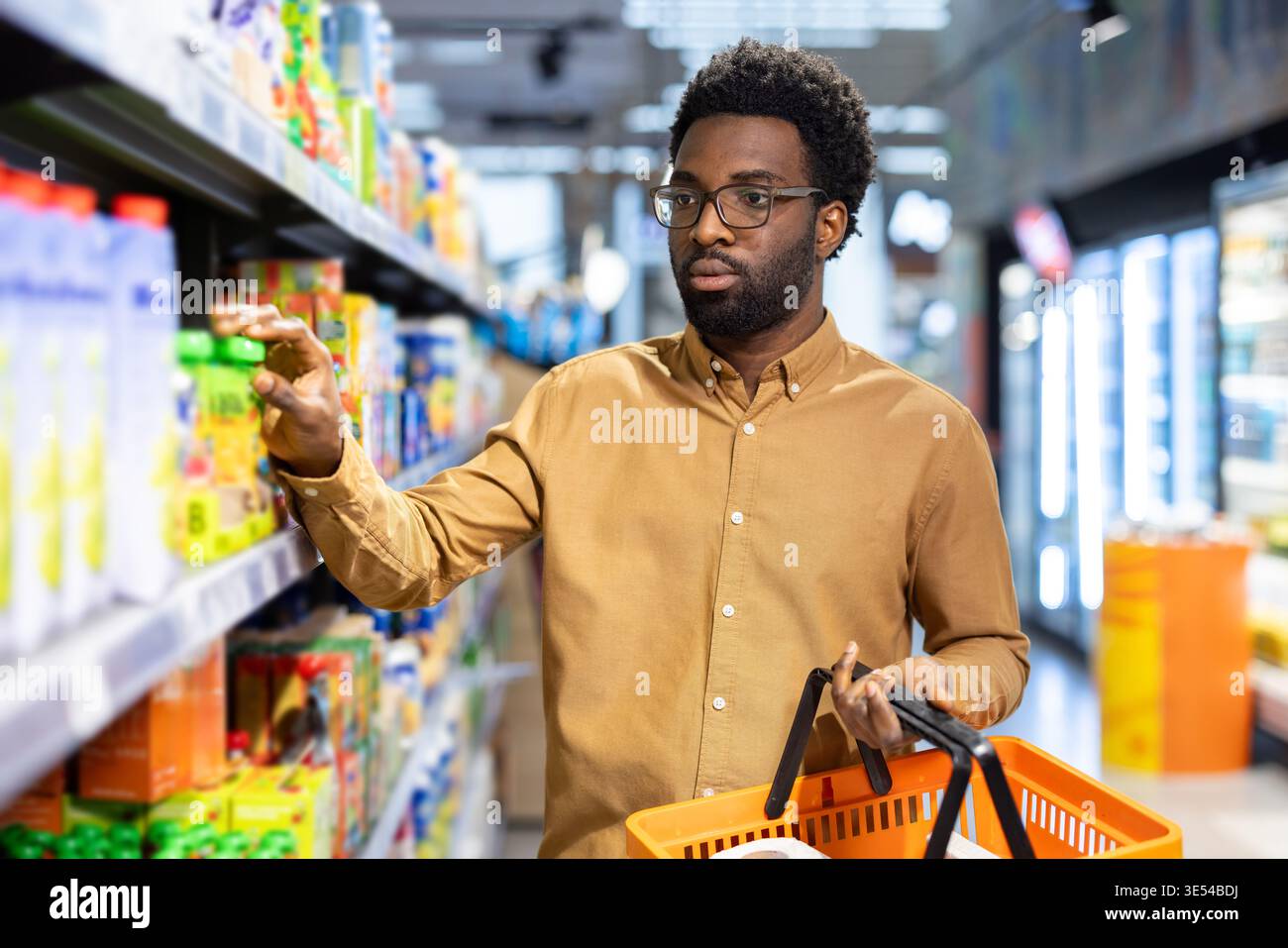 Homme afro-américain avec des lunettes et un panier examinant soigneusement les produits sur une étagère bien éclairée dans une épicerie moderne, prenant des décisions d'achat Banque D'Images