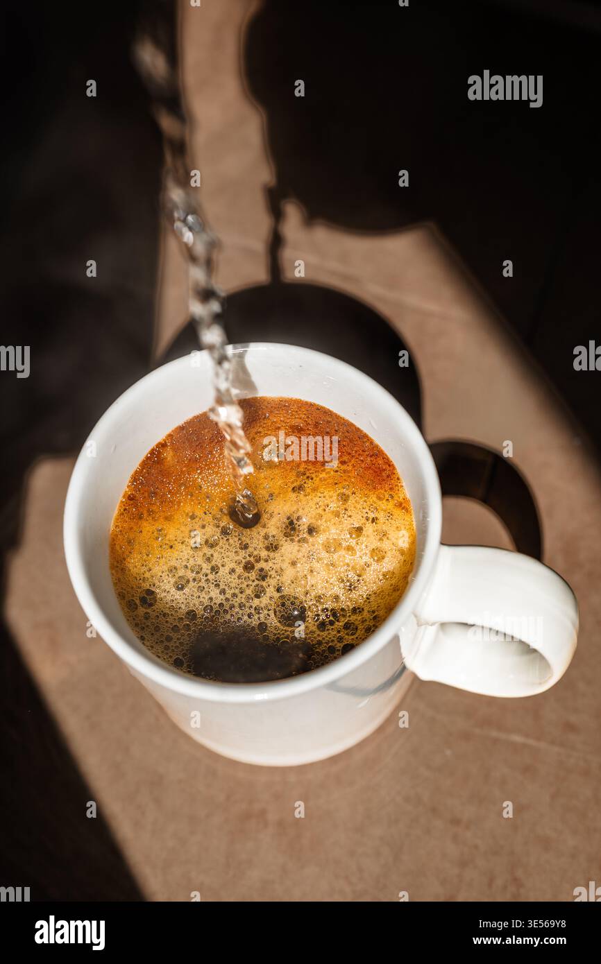 De l'eau chaude fraîche étant versée dans une tasse en céramique blanche de café instantané avec de la mousse dorée. Banque D'Images