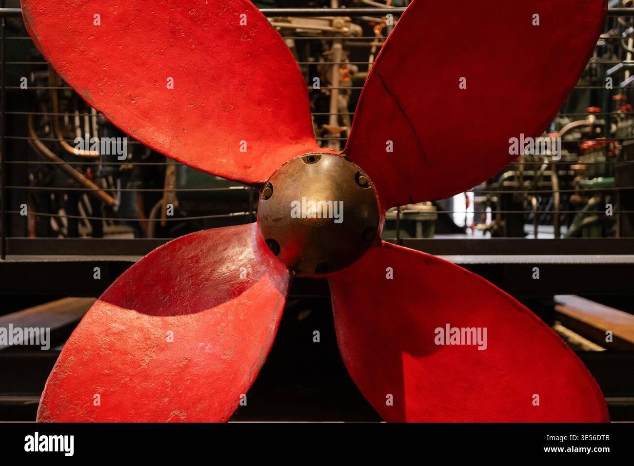 Hélice de navire rouge vintage. Une grande hélice de navire à quatre pales altérée peinte en rouge vif avec un moyeu en laiton, exposée contre des machines industrielles. Banque D'Images