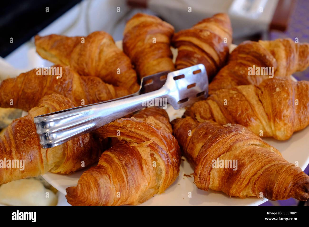 Croissants simples fraîchement cuits disposés en grappe, pinces posées sur un élégant plat en porcelaine pourpre. Sopot. Pologne. Banque D'Images