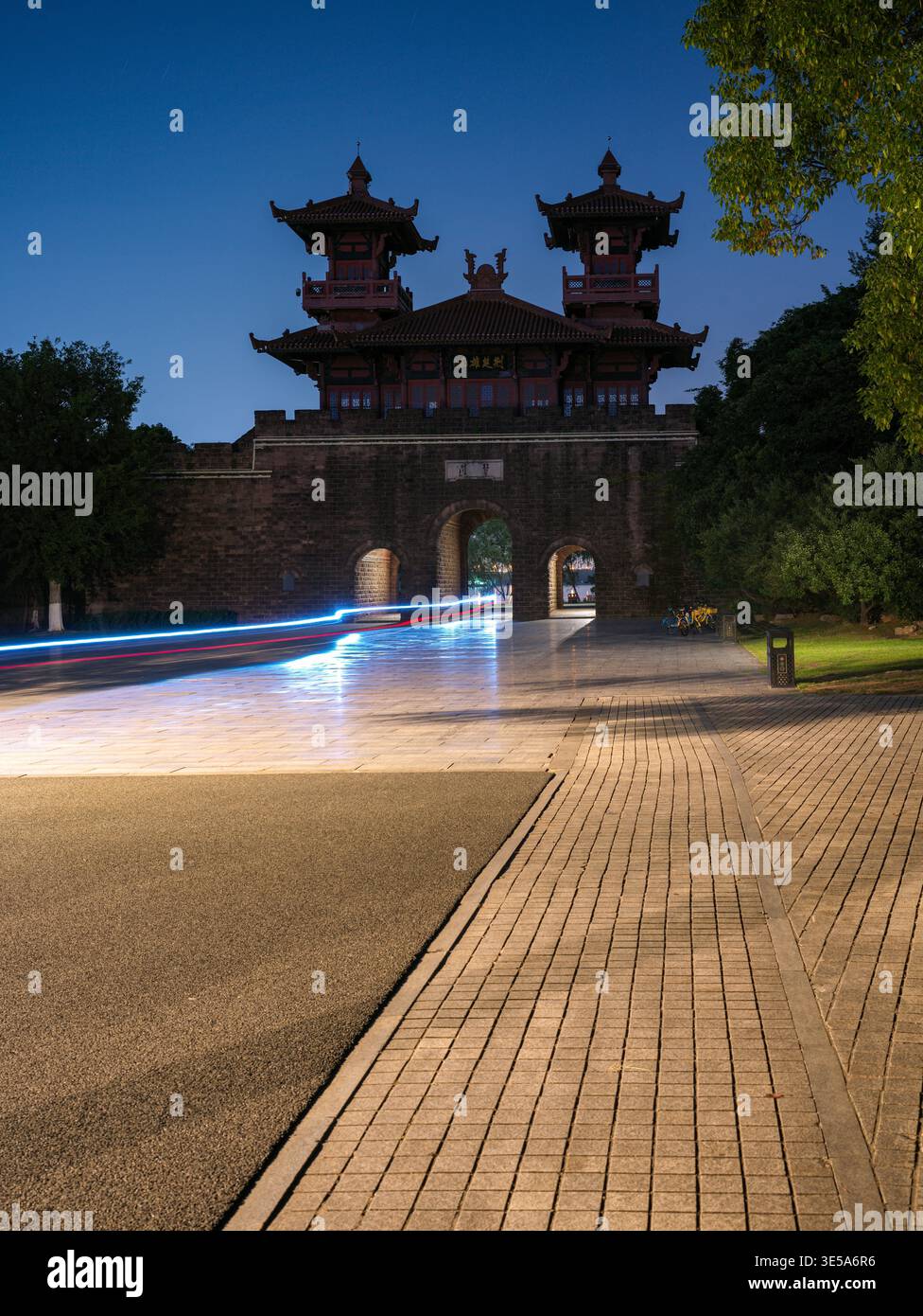 La porte d'entrée traditionnelle chinoise historique en brique illuminée contre un ciel nocturne sombre, avec des traînées de lumière rouge et blanche floues passant rapidement dans le Banque D'Images