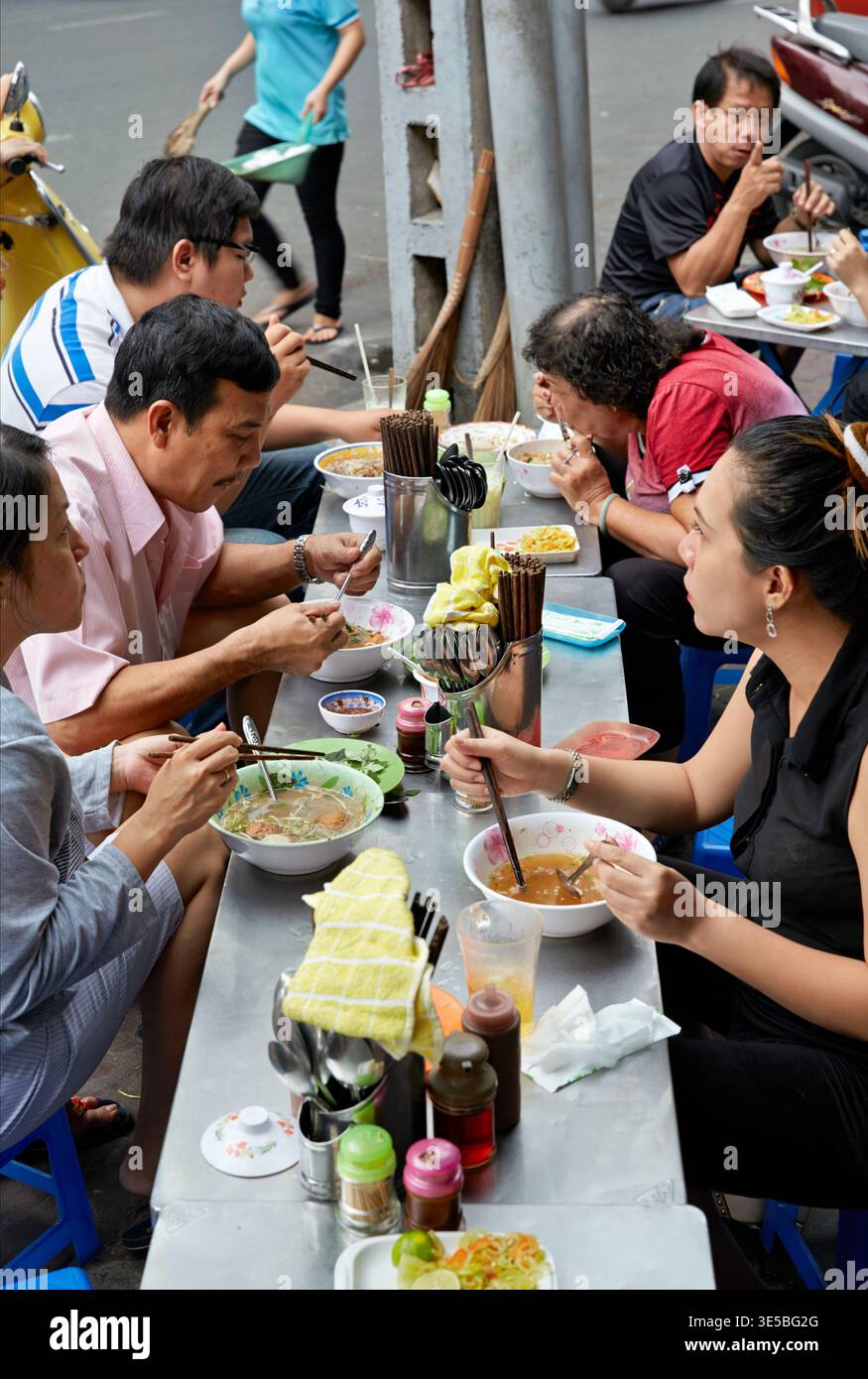 Des groupes de personnes apprécient des bols de nourriture dans les étals de rue à Saigon, au Vietnam. Ils utilisent des baguettes pour manger divers plats dans un cadre extérieur. Banque D'Images