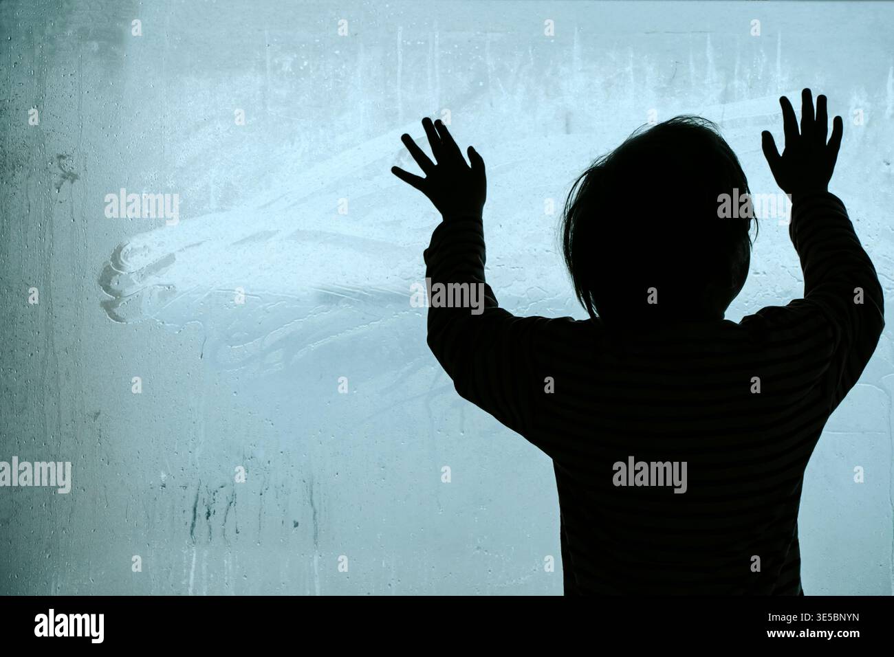 Silhouette d'un enfant solitaire regardant à travers une grande fenêtre embuée froide. Un travail de photographie conceptuelle explorant les thèmes de la dépression infantile, de la crise psychologique et du profond sentiment d’isolement et de tristesse. Photo de haute qualité Banque D'Images